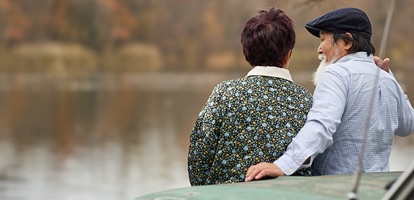 A man with a blue hat holding his left arm around a woman as they sit and facing away towards a lake.