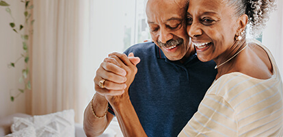 An older couple smiling as they dance cheek to cheek in their living room 