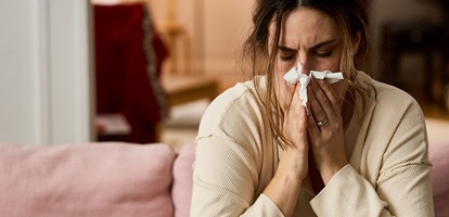An unwell woman sitting on a couch and blowing her nose with a tissue. 