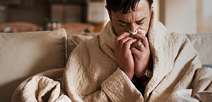 An unwell man sitting on a couch and blowing his nose with a tissue. 